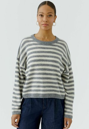 Frau trägt einen grau-weiß gestreiften Pullover, blaue Jeans und goldene Creolen, steht vor einem schlichten hellen Hintergrund.