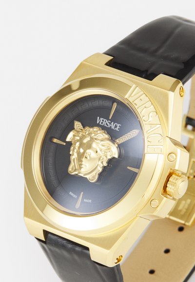 Auksinės spalvos laikrodis su juodu odiniu dirželiu, turintis juodą ciferblatą, iškilų Medūzos emblemą ir įrašytą „VERSACE“ ant rėmelio.