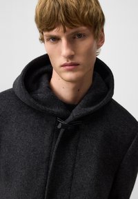 Donkergrijze wollen jas met een capuchon, voorzien van een toggelsluiting en een zachte textuur. Eenvoudig ontwerp zonder zichtbare patronen of accenten.