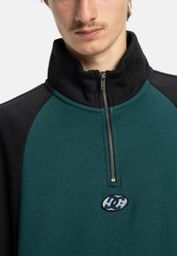 Pulôver com meio zíper em verde escuro e preto, feito de tecido suave. Apresenta um patch de logotipo e um fecho de correr suave ao longo do colarinho.