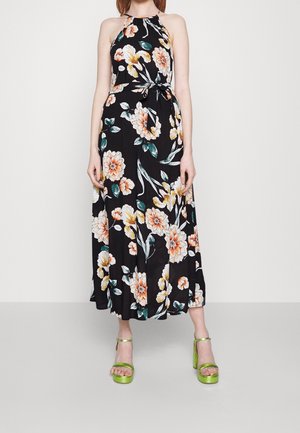 Femme portant une robe noire sans manches à fleurs avec un col halter et une ceinture à la taille, ainsi que des sandales vertes à plateforme à talons hauts, debout devant un fond uni.