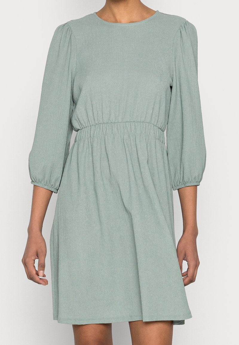 Femme portant une robe texturée vert clair avec de longues manches bouffantes et une taille élastiquée, debout devant un fond uni.