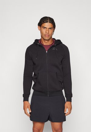 Moški z pletenimi lasmi, oblečen v črn pulover z zadrgo čez bordo majico in črne športne kratke hlače, stoji na enostavni ozadju.