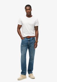 Camiseta blanca de cuello redondo, jeans azules, cinturón de cuero marrón, zapatillas crema, pose de pie. Diseño simple, corte casual.