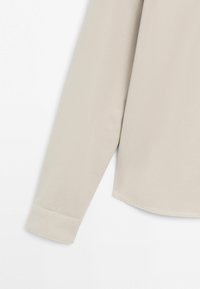 Beige langärmliges Hemd mit Manschettendetail vor weißem Hintergrund, das glatte Stoffstruktur und saubere Nähte zeigt.