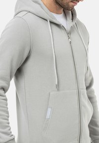 Haut de sweatshirt zippé gris clair avec capuche, doté d'une poche avant, de cordons de serrage et d'une fermeture éclair argentée. Fabriqué en tissu doux et texturé.