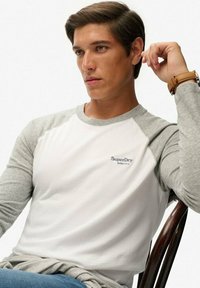Koszulka z długim rękawem typu raglan z białym tułowiem i szarymi rękawami. Posiada małe logo "SuperDry" na klatce piersiowej. Miękka tkanina w składzie bawełny.
