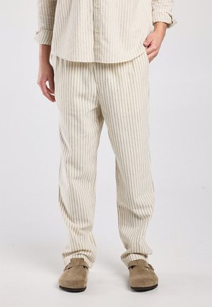 Persoon die beige broek met krijtstreepjes en bijpassend overhemd draagt, met beige suède instapschoenen, staande met één hand in de zak.