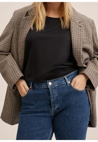 Blazer à motif pied-de-poule marron et noir porté sur un haut noir, associé à un jean en denim bleu. Le jean est doté d'une poche avant et d'une fermeture à bouton.