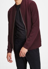 Burgundy bomber jacket with a silver zipper, ribbed cuffs, and a smooth fabric texture. Worn over a black shirt, paired with black pants.