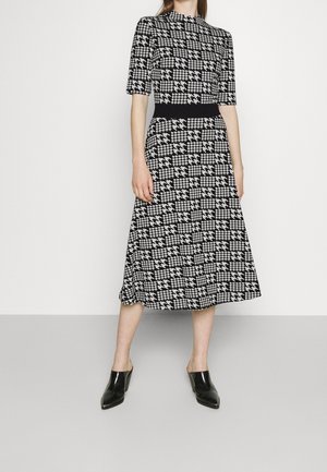 Frau trägt ein schwarz-weißes Hahnentritt-Midikleid mit halblangen Ärmeln und einem schwarzen Taillenband, kombiniert mit schwarzen spitzen Slippern.