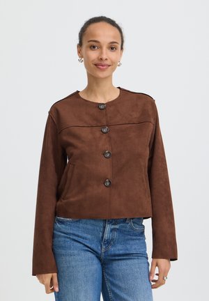 Femme aux cheveux bouclés portant une veste en daim marron boutonnée et un jean bleu, debout devant un fond clair uni.