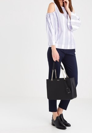 Femme portant un chemisier rayé à épaules dénudées, pantalon court marine, bottines noires, tenant un sac à main en cuir noir avec des anses en chaîne dorée.