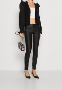 Svart cardigan med spetsiga puffärmar, vit crop top, svarta skinnbyxor i skinny-modell, svarta spetsiga klackskor och en rosa mönstrad handväska.