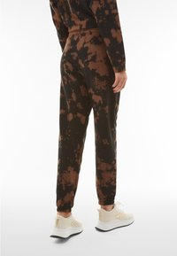 Black joggers with brown tie-dye pattern, elastic waistband, and cuffed ankles, paired with light-colored sneakers featuring a textured design.
