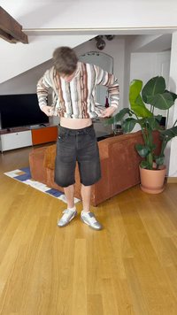 Πολύχρωμο ριγέ πουκάμισο με μακρύ μανίκι, μαύρες τζιν βερμούδες και ασημένια αθλητικά παπούτσια. Ξύλινο δάπεδο, πράσινο φυτό και πορτοκαλί καναπές στο φόντο.