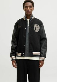 Ung mand iført en sort varsity-jakke med læderærmer, broderede logoer, lagdelt over en grå zip-up sweater og en hvid skjorte.