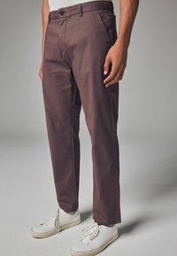 Brune bomuldsbukser med lige pasform, forlommer og knaplukning, parret med hvide sneakers med et struktureret design.
