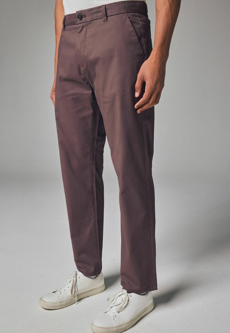 Brune bomuldsbukser med lige pasform, forlommer og knaplukning, parret med hvide sneakers med et struktureret design.