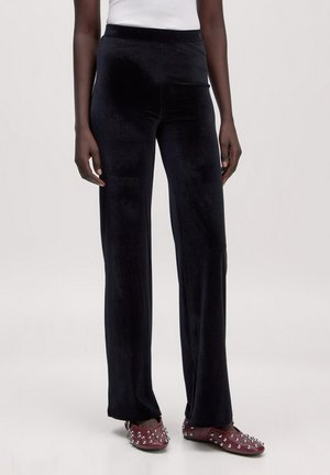 Frau trägt schwarze Velvet-Weitbein-Hosen kombiniert mit burgunderfarbenen, mit Nieten besetzten Mary Jane Ballerinas und einem weißen ärmellosen Oberteil.