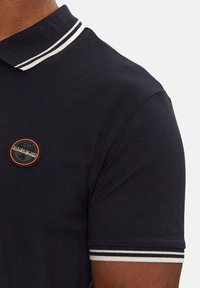 Polo shirt blu navy con strisce bianche sul colletto e sulle maniche. Presenta un logo circolare con dettagli neri e arancioni sul petto.