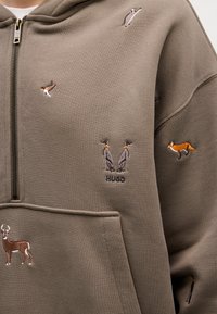 Sudadera verde oliva hecha de algodón, con bordados de varios animales: un ciervo, galgos, un zorro y golondrinas. Con cremallera en la parte delantera y bolsillo canguro.