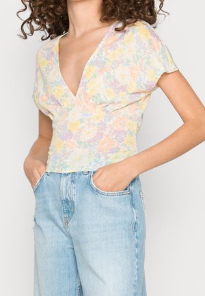 Femme aux cheveux bouclés portant un chemisier pastel à fleurs avec col en V et un jean taille haute bleu clair, les mains dans les poches, sur fond uni.