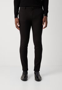 Zwarte slim-fit broeken met een gladde textuur, voorzien van een ritssluiting en een enkele knoopsluiting, gecombineerd met zwarte leren schoenen.