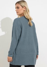 Un pull à manches longues, ample, en tricot bleu doux et texturé. Présente des fentes sur les côtés et des détails perforés le long des manches.