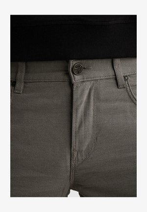 Gros plan sur un pantalon gris texturé avec un bouton visible et des passants de ceinture, porté avec un haut noir côtelé.
