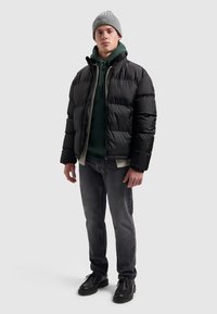 Pufferjack in zwart, voorzien van een rits- en drukknopsluiting, gedragen over een groene hoodie, gecombineerd met grijze jeans en zwarte schoenen. Grijze gebreide beanie.
