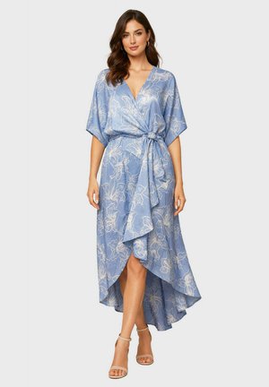 Femme portant une robe portefeuille fleurie bleu clair avec un ourlet asymétrique et des manches courtes, associée à des escarpins nude à bride cheville.