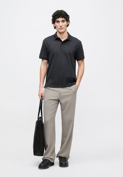 Melnas polo krekls ar faktūru zigzaga rakstu, kombinēts ar gaiši pelēkām biksēm. Tur melnu somu un valkā melnas kurpes.