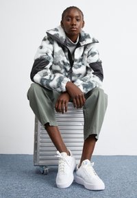 Fluffig vit och gråmönstrad jacka med svarta ärmar, olivgröna byxor och vita sneakers, sittande på en silverfärgad resväska.