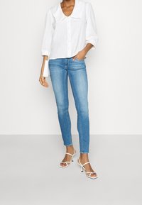 Kvinna som bär ljusblå skinny jeans, vit skjorta med stor krage, vita klacksandaler och bär en liten vit axelväska.