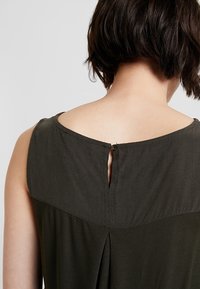 Robe sans manches vert foncé avec un col rond, une ouverture en trou de serrure au dos avec une fermeture à bouton, et une texture subtile sur le tissu.