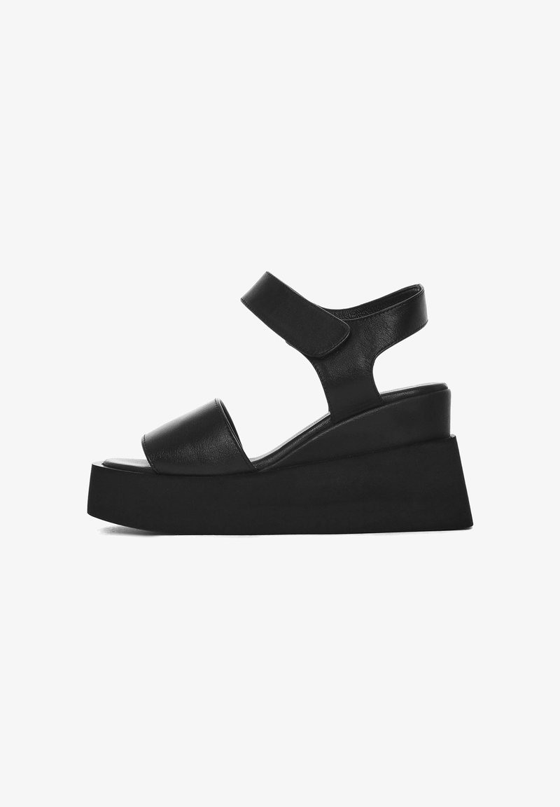 Sandales à talon compensé noires avec une tige en cuir lisse, une sangle adjustable, un bout ouvert et une semelle épaisse. Design simple sans embellissements.