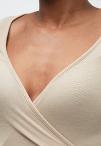 Bēša ribota auduma augšdaļa ar dziļu V veida kakla izgriezumu. Tekstūra ir gluda, ar redzamām šuvēm gar kakla izgriezumu un apakšu.