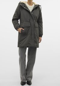 Oliwkowo-zielona pikowana parka z kapturem wyłożonym sztucznym futrem, regulowanym sznurkiem w talii i dwoma przednimi kieszeniami; w zestawie z luźnymi szarymi spodniami.