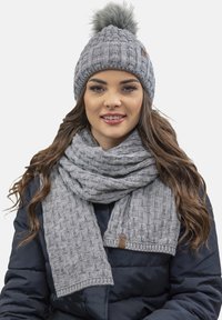 Bonnet en tricot gris avec un pompon en fausse fourrure, associé à une écharpe à côtes assortie. Les deux sont fabriqués dans un tissu doux et texturé.