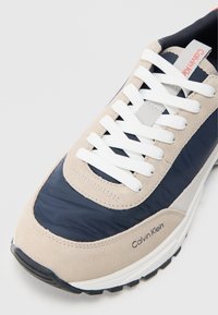 Tennised, millel on beeži ja tumesinine disain, sametmaterjal ja võrgusilma, valged paelad ning must kummist tallaga, millel on "Calvin Klein" brändimine.