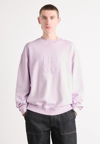 Gaiši violets džemperis ar ribotu apkakli un elastīgiem piedurknēm. Priekšpusē ir izšūts "HUGO" logo. Valkāts kopā ar tumšiem džinsu biksēm.