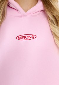 Felpa rosa realizzata in morbido tessuto, con tasca frontale e un logo ricamato rosso "MIRONS" all'interno di una forma ovale.