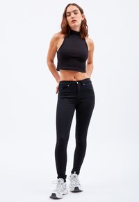 Schwarzes Cropped Top mit hohem Kragen, kombiniert mit schwarzen Skinny Jeans. Getragen mit weißen Sneakers. Glatte Textur, enganliegendes Design.