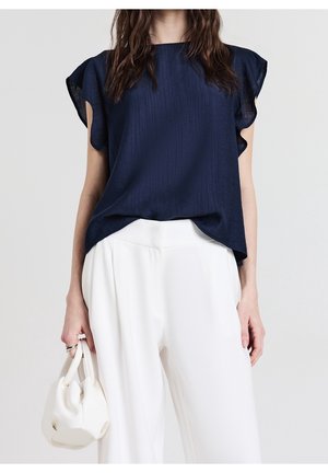 Femme portant un chemisier bleu marine à manches volantées et un pantalon blanc à jambes larges, tenant un petit sac à main blanc dans sa main gauche.