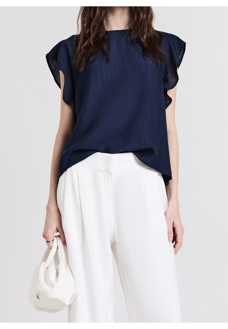 Femme portant un chemisier bleu marine à manches volantées et un pantalon blanc à jambes larges, tenant un petit sac à main blanc dans sa main gauche.