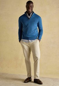 Zils džemperis ar rāvējslēdzēja apkakli, valkāts pār rūtainu krekliņu ar apkakli, bēšas krāsas bikses un brūnas kurpes bez šņorēm. Gluda tekstūra un pieguļošs piegriezums. Neitrāla fona krāsa.