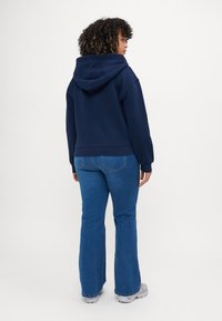 Mujer con cabello rizado que lleva una sudadera azul marino, jeans azules y zapatillas blancas, de pie contra un fondo blanco liso.
