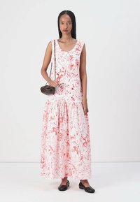 Weißes Maxikleid mit rotem abstraktem Blumenmuster, ärmelloses Design, taillierter Oberteil und ausgestellt, gestuften Rock. Dunkle Handtasche und schwarze Ballerinas.