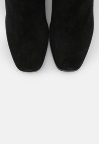 Botas de tobillo de ante negras con una textura suave, diseño de puntera cuadrada y detalles minimalistas, sobre un fondo blanco.